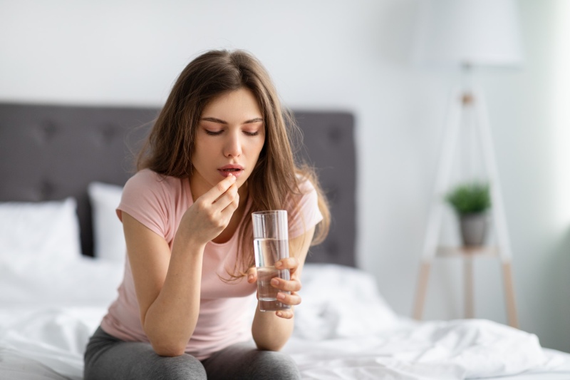 Young Caucasian lady holding glass of water, taking pill on bed, feeling unwell at home, copy space. Millennial woman using medical drug, suffering from headache, drinking tablet indoors